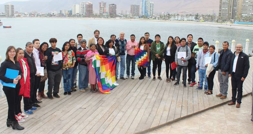 A diverse group posing with a colorful flag by the waterfront, with city buildings in the background.